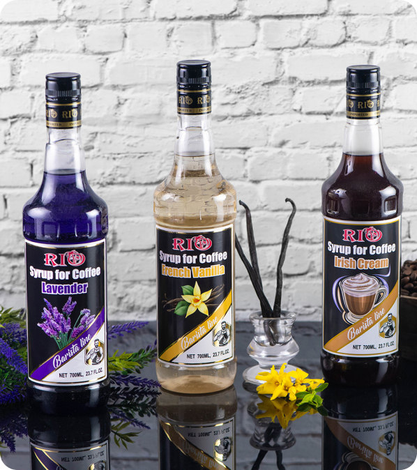 Three bottles are displayed against a white brick wall. The flavours are Lavender, French Vanilla, and Irish Cream. A sprig of lavender, vanilla beans, a yellow flower, coffee beans, and coffee drinks surrounds them.