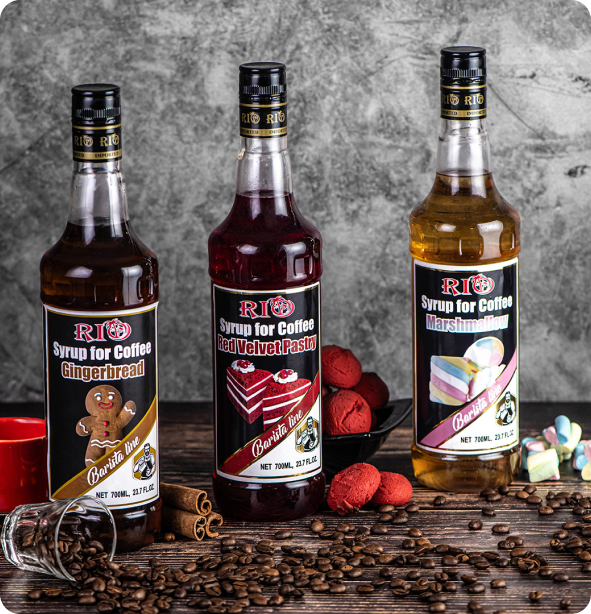 Three bottles of Rio Syrup for Coffee stand on a table surrounded by coffee beans, red macarons, and marshmallows. The flavors displayed are Gingerbread, Red Velvet Pastry, and Marshmallow.