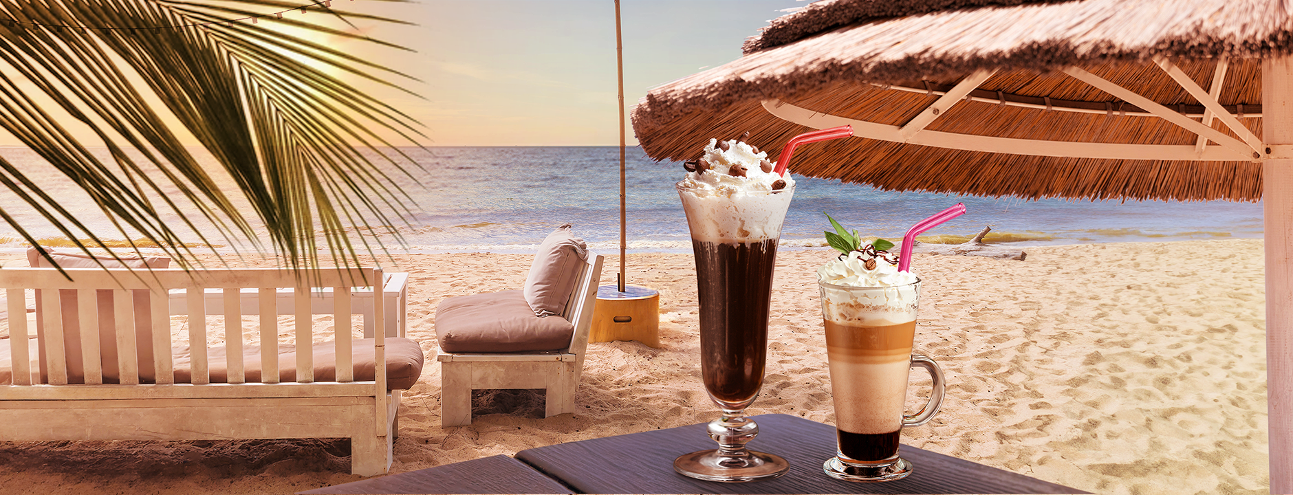 Two iced coffee drinks in tall glasses with whipped cream and straws are on a table at a sandy beach. A straw parasol and lounge chairs are in the background, with a view of the blue sea and a serene sunset.