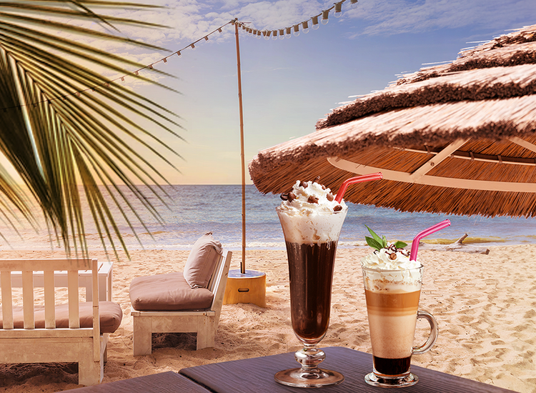 Two iced coffee beverages topped with whipped cream, chocolate shavings, and cherries are on a table under a straw umbrella at a beach.