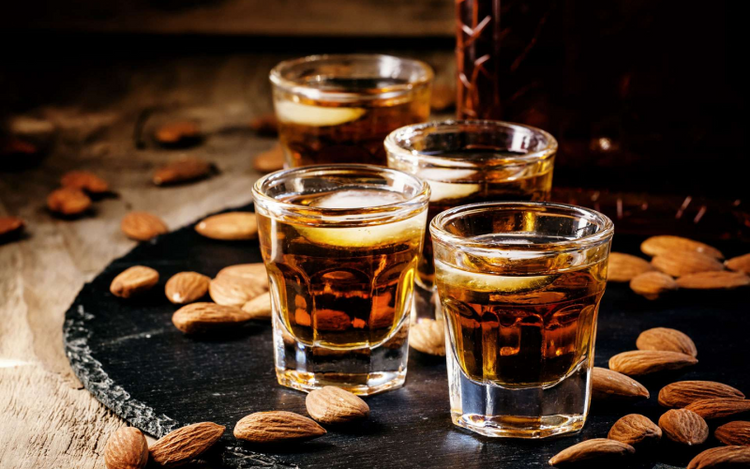 Four shot glasses filled with a golden-brown liquid are placed on a rustic black slate surface. Almonds are scattered around the drinks.