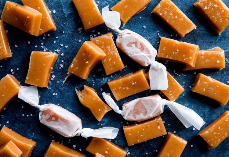 Rectangular pieces of caramel candy, some wrapped in white paper, scattered on a dark surface with sprinkled sea salt crystals.