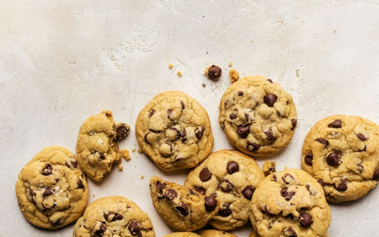 chocolate chip cookies scattered on a light beige surface. Some cookies have crumbs around them, and the cookies are golden-brown with visible chocolate chips embedded in them.