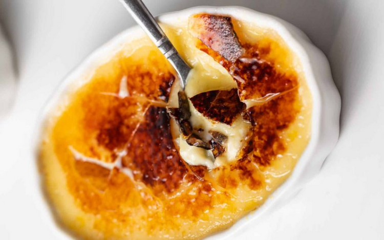 A close-up shot of a crème brûlée in a white ramekin. The caramelized sugar top has been cracked with a silver spoon, revealing the creamy custard underneath.