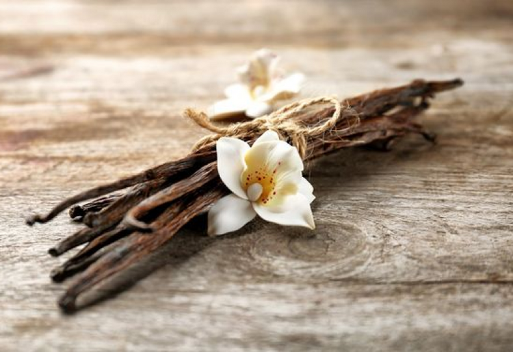 A bundle of dried vanilla beans tied together with twine lies on a wooden surface. Two white vanilla flowers are placed on top of the bundle