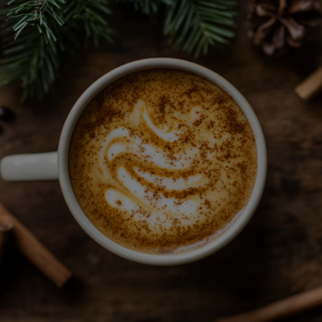 A cup of frothy coffee with latte art, sprinkled with cinnamon, sits on a wooden surface. Surrounding the cup are pine branches, a cinnamon stick, and a pinecone, creating a cozy, festive atmosphere.