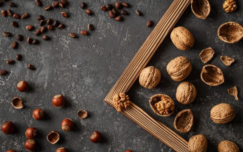 Various whole and cracked walnuts and hazelnuts scattered on a dark textured surface. A wooden frame is positioned diagonally, partially enclosing some nuts.