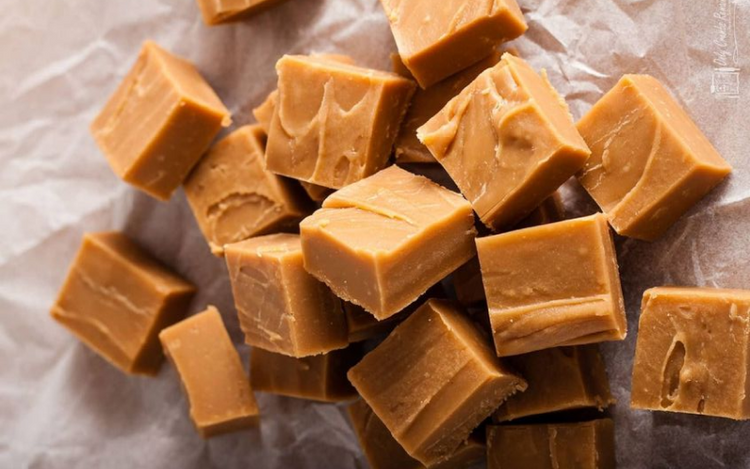 A collection of fudge squares is spread out on a piece of crumpled parchment paper. The fudge has a smooth, creamy texture and a rich caramel color. The squares are neatly cut and stacked randomly.