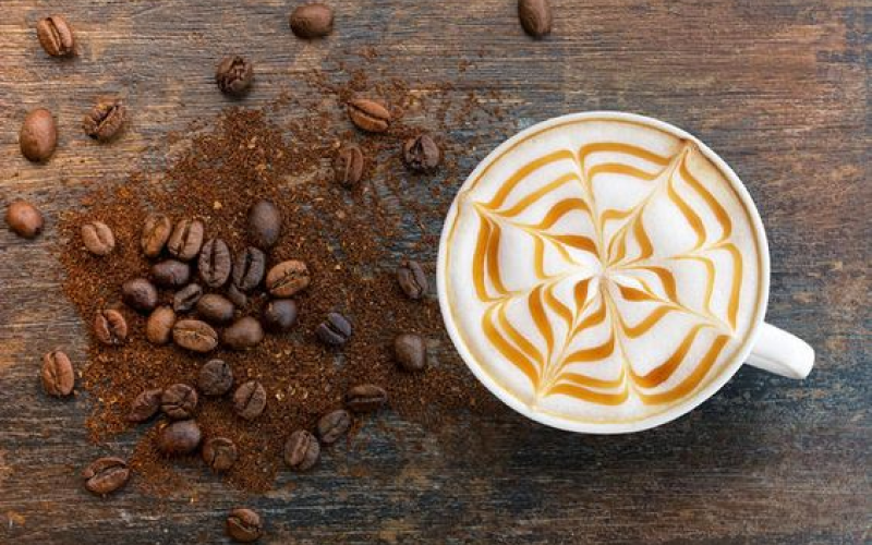 A cup of coffee with intricate caramel latte art on top sits on a rustic wooden surface. Next to the cup, there are scattered coffee beans and ground coffee,