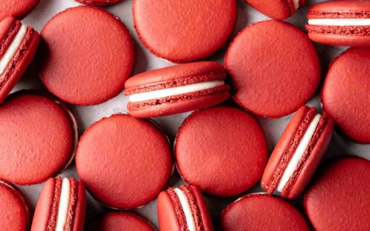 Numerous red macarons. Some of the macarons are whole, while others are sandwiched together with a white cream filling visible in the middle. The macarons are arranged closely together, showcasing their vibrant red color and smooth texture.